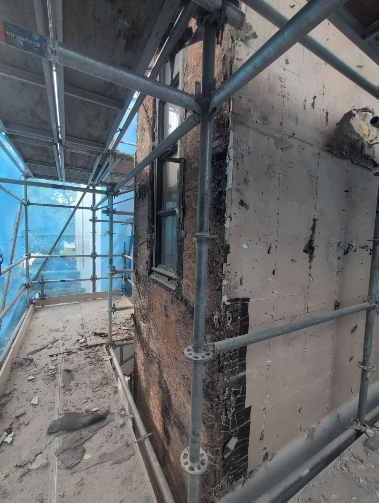 Exterior wall stripped back to sheathing during stucco demolition, scaffold in place, black water staining across the substrate