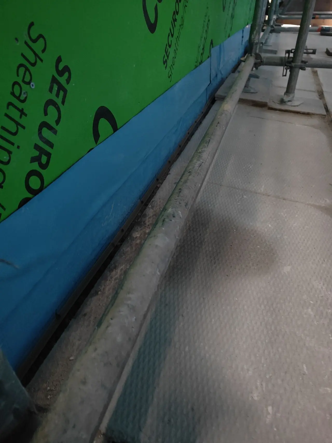 Blue self-adhered waterproofing membrane lapped from exterior sheathing down over the wall base and foundation, with the base flashing termination below