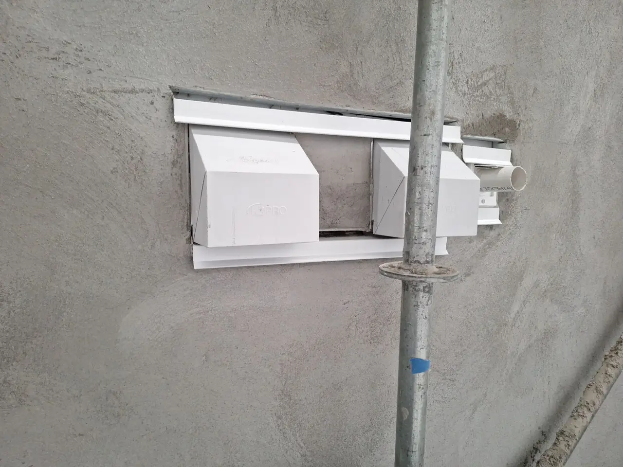 Two white hood vents and a pipe penetration set in metal trim against a finished Portland cement brown coat, showing the open drainage gap above the head trim