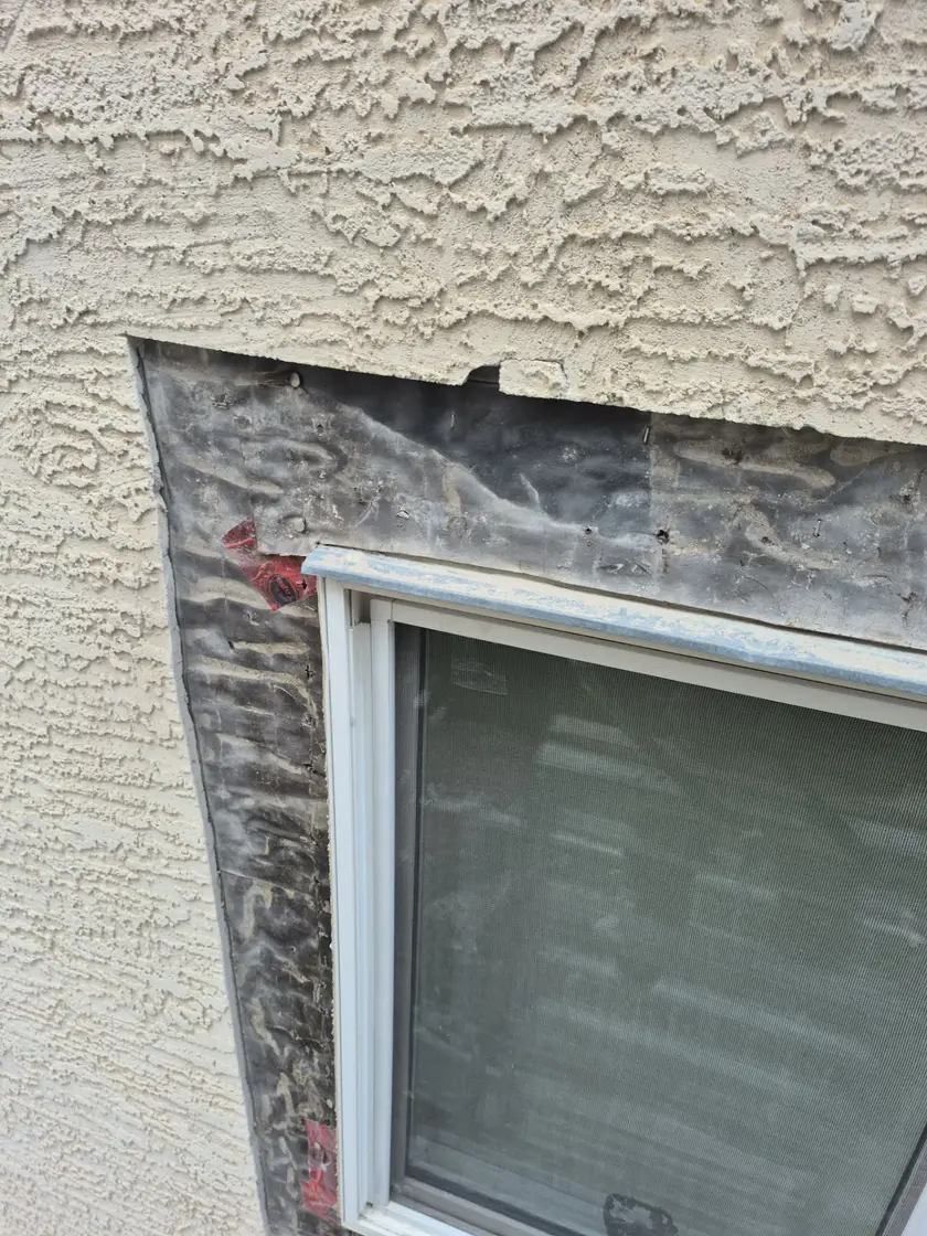 Stucco cut back to a straight square edge around a window opening, exposing existing building paper and window nail flange